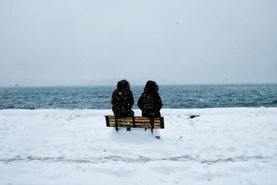Rear view of friends sitting on bench at beach during snowfall