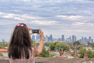 Rear view of woman photographing buildings using phone against cloudy sky