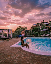 Full length of couple standing by swimming pool against sky during sunset