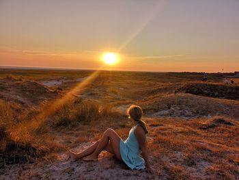 Side view of woman sitting on field against sky during sunset