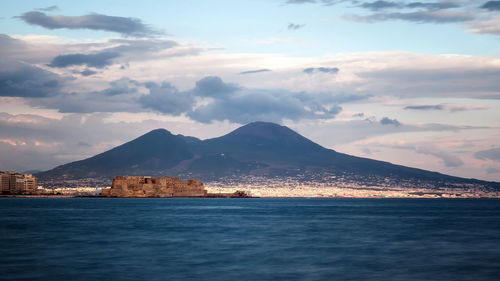 Mid distance view of castel dellovo by sea with mountains in background against cloudy sky
