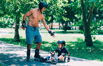 Shirtless grandfather giving hand to grandson fallen on footpath while skating in park
