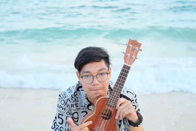 High angle portrait of young man holding violin while sitting at beach during sunset