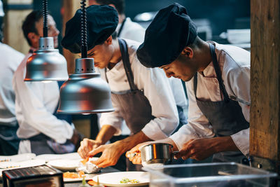 Men in hats and aprons working in team and serving dishes with delicious food in kitchen