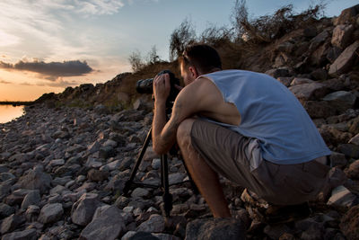 Side view of man photographing at beach against sky during sunset