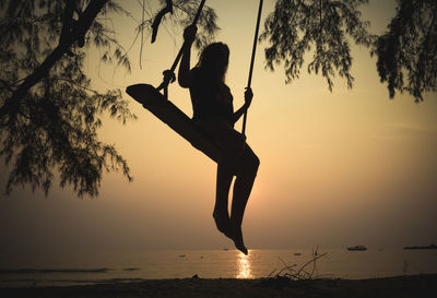 Silhouette girl swinging at beach against sky during sunset