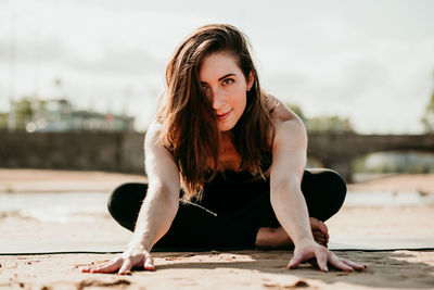 Smiling flexible female sitting on mat in janu sirsasana and doing forward bend while looking at camera and practicing yoga on beach in summer