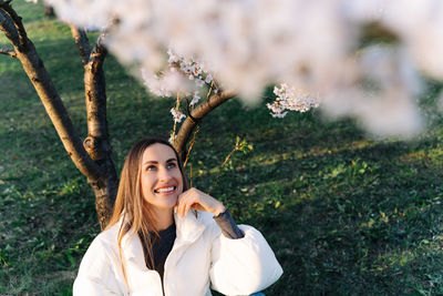 Beautiful woman in a white jacket sits under blooming tree in city park in spring