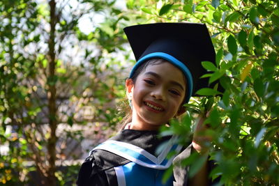 Portrait of smiling girl wearing graduation gown and mortarboard by plants