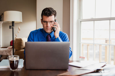 Man on cellphone working from home using a computer at a dining table.