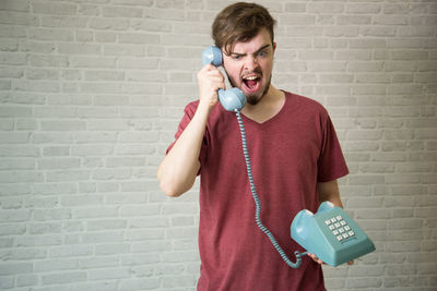 Young man yawning while using telephone against brick wall