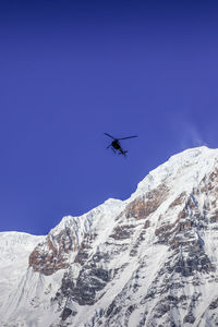 Low angle view of helicopter flying over snowcapped mountains against clear blue sky