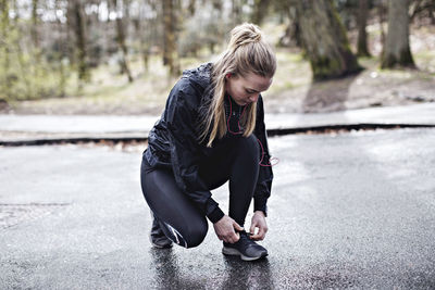 Full length of female athlete tying shoelace while crouching on wet road in forest