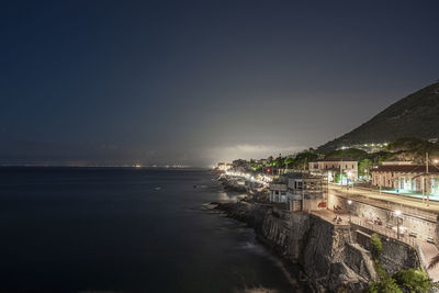 Illuminated buildings by sea against sky at night