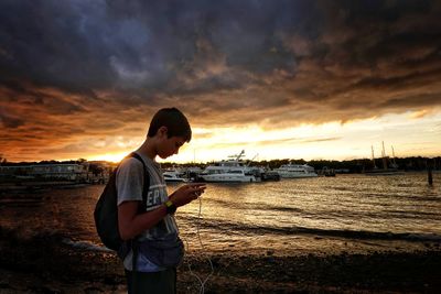 Side view of boy standing at beach against cloudy sky during sunset