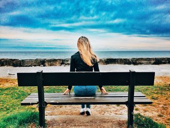 Rear view of man sitting on beach against cloudy sky