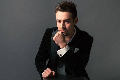 Portrait of arrogant young man in suit sitting with hand on chin against gray background