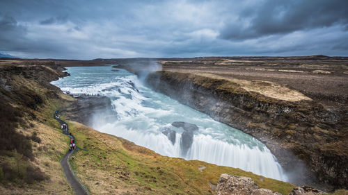 People walking on footpath by beautiful waterfall against cloudy sky