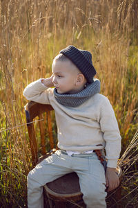 Boy in warm clothes sit on chair along a path on a field with dried grass in autumn