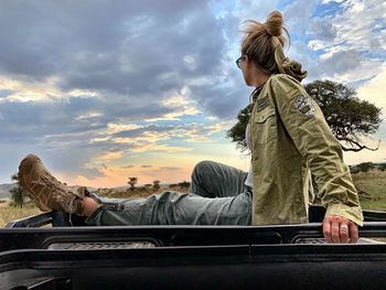 Side view of woman sitting on car roof against sky during sunset