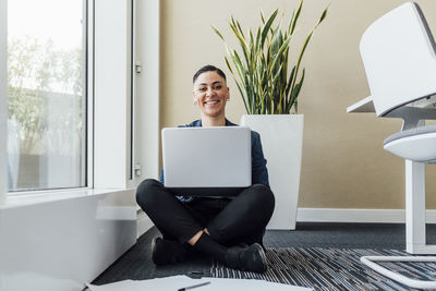 Smiling businesswoman with laptop on lap sitting on floor in office