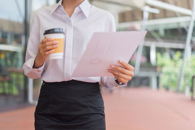 Midsection of businesswoman holding disposable cup and papers while standing outdoors
