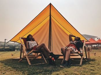 Women relaxing on chairs under tent at campsite against clear sky