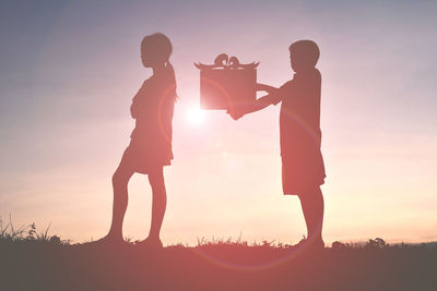 Silhouette boy giving gift to girl on field against sky during sunset