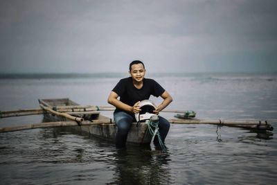 Full length portrait of young man sitting in sea against sky