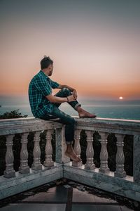 Full length of man looking at view while sitting on railing against sky during sunset