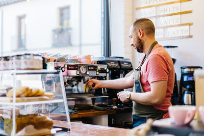 Side view of male barista in apron with portafilter brewing coffee using coffee machine while standing near counter in coffee house