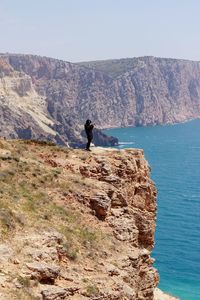 Rear view of human with camera standing in rocky mountains against blue sea 