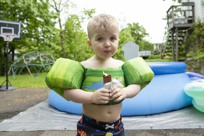 Young boy wearing swimming gear stands eating candy in yard with pool