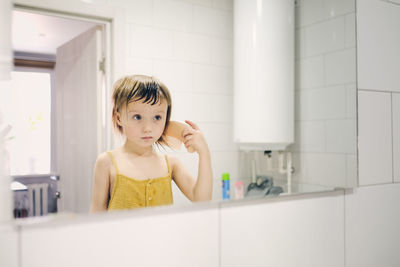 Child in yellow jumpsuit combs his own in front of mirror in bathroom