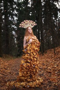 Thoughtful young woman covered with leaves standing at forest during autumn