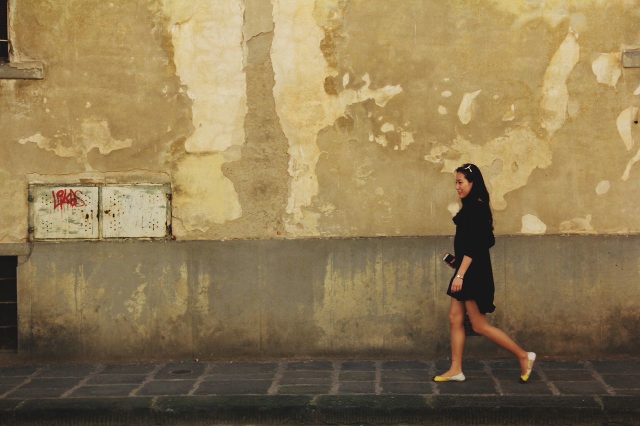 wall - building feature, lifestyles, full length, architecture, casual clothing, built structure, young adult, standing, leisure activity, wall, person, brick wall, building exterior, young women, indoors, side view, front view, three quarter length