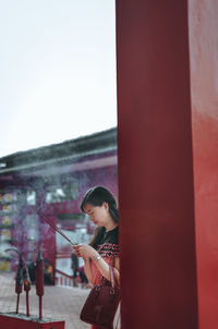 Side view of young woman holding red umbrella against built structure