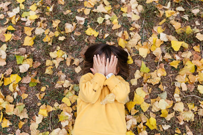 Upper view on little girl hiding face with hands lying between yellow leaves