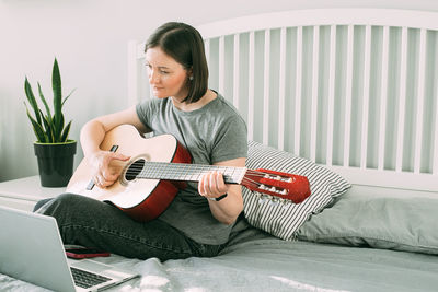 Young man playing guitar while sitting on bed at home