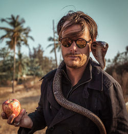 Portrait of mature man with snake holding apple standing against sky