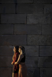 Two girls standing against stone wall during sunset
