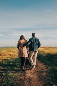 Back view of tender couple hugging and walking with baby stroller along road in countryside on sunny day