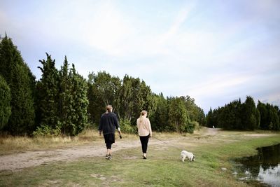 Rear view of two friends walking around with small white dog on a path surrounded by trees and lake 