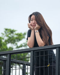 Young woman smiling while standing by railing against sky