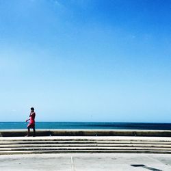 Man walking on side walk by sea against clear blue sky