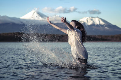 Young woman splashing water while standing in lake against snowcapped mountain