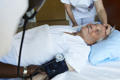 Male nurse checking blood pressure of senior man on hospital bed while colleague consoling patient