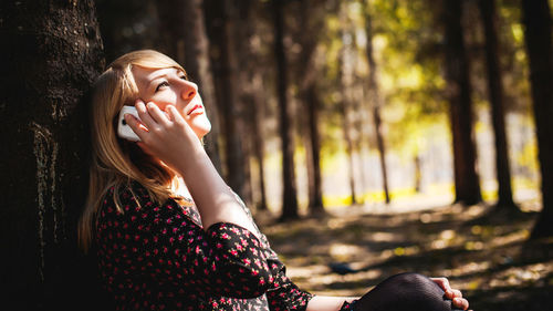 Woman talking on mobile phone while sitting by tree trunk in forest