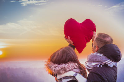 Young couple holding red heart shape cushion against sky during sunset
