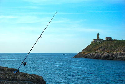 Fishing rod on rock by sea against blue sky